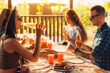 Mutlu bir arkadaş grubu arka bahçe partisinde eğleniyor gençler birlikte gülüyor, bira içiyor ve dışarıda yemek yiyor.