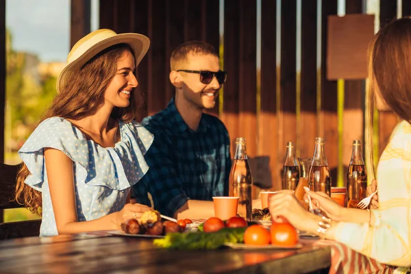 Gençler terasta eğleniyor, içki içiyor ve sohbet ederken arkadaşları gün batımında dışarıda ızgara et servis ediyor.