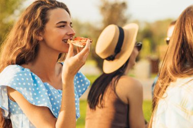 Yaz pikniği, üç gülümseyen güzel kız arkadaş, çimenlerde oturmak, güneşli bir yaz gününde pizza yemek ve içki içmek.