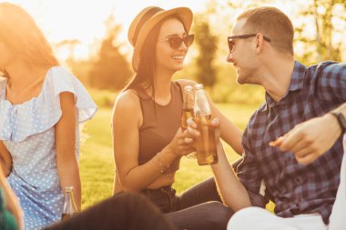 Yazlık şapkalı ve güneş gözlüklü güzel bir kadın güneşli bir günde parkta piknikte içki içen genç bir adamla iletişim kuruyor.