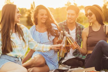 Bir grup öğrenci dışarıda piknikte yemek yiyor, bir sürü lezzetli yemek yiyor, pizza yiyor ve soğuk içecekler içiyor, çimlerin üzerinde, güneşli bir havada, battaniyede birlikte oturuyorlar.