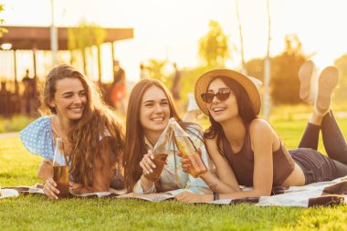 Üç güzel gülümseyen kız arkadaş, kız arkadaşlar soğuk içecekler içip sohbet ediyorlar, güneşli bir günde yeşil bir çimenlikte güneşleniyorlar.
