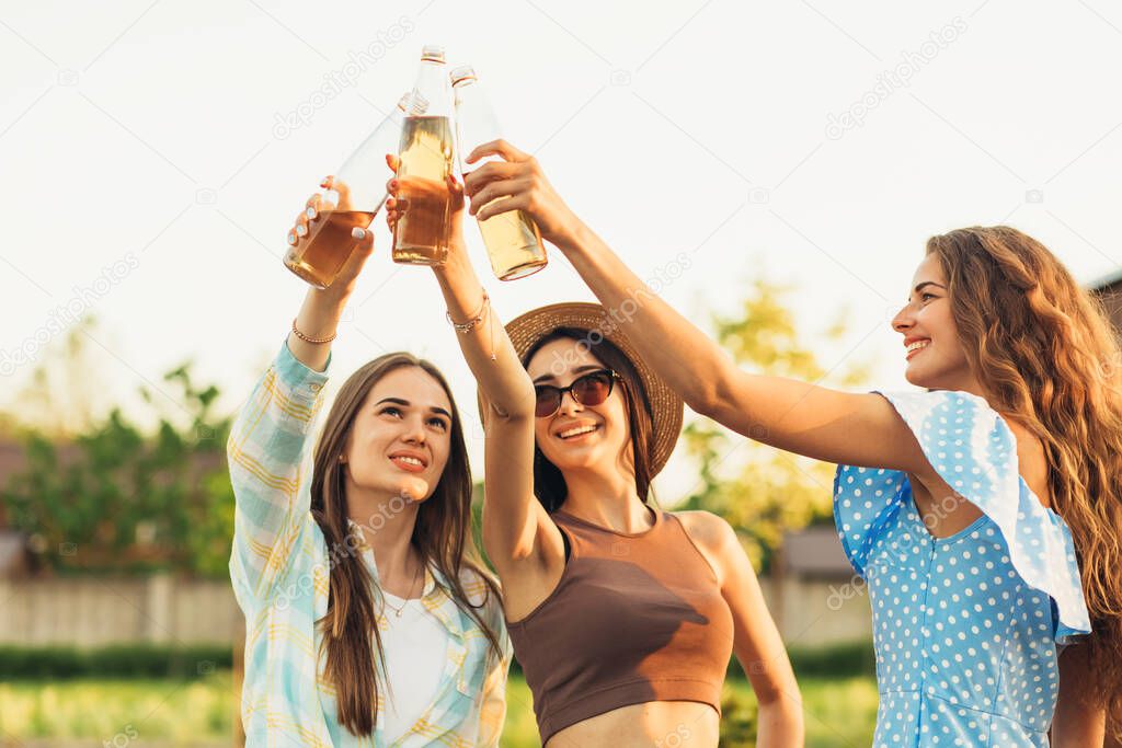 Three beautiful smiling young women friends walking in the park, celebrating and drinking beer drinks, outdoors at sunset, victory and friendship concept