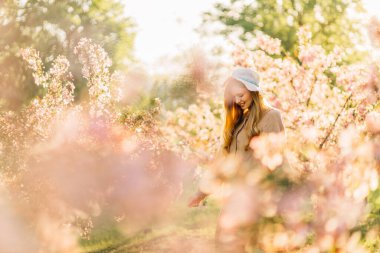 Sarı saçlı, narin elbiseli, bahar bahçesinde çiçek açan kiraz ağaçları olan güzel, genç bir kadın.