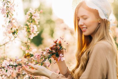 Güzel bahar genç bayanı şapkalı, güzel kokulu pembe çiçeklerle çiçeklerle bir yaz parkında, çiçek açan bir bahçede, güneşli bir bahar gününde, güzellik konsepti, doğallık