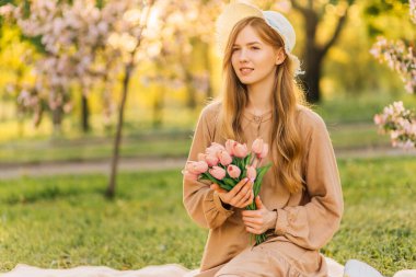 Bir buket bahar çiçeğiyle gülümseyen güzel bir kadın, hasır bir yaz şapkasıyla, baharda bir bahçede çimenlikte piknik yapan bir kadın, gün batımında çiçek açan bir bahçede.