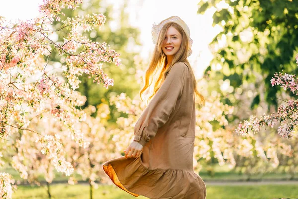Yazlık şapkalı güzel bir kadın ve çiçek açan bahçelerde dans eden bej renkli bir elbise baharda çiçek açan ağaçların altında mutlu bir kadın.