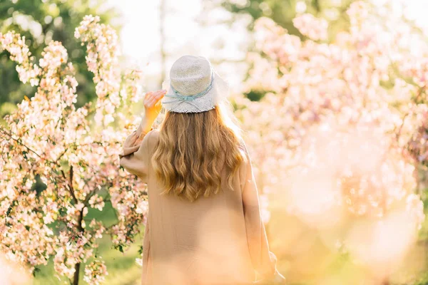 Şapkalı hoş bir genç kadın, narin bir elbise giymiş, ağaçların renkli pembe çiçeklerinin yanında duruyor, romantik kadınlar.