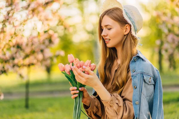 美丽的年轻女子头戴一顶帽子 捧着一束春花 在春天里的樱花花园里 花瓣从树上掉下来 春意盎然 — 图库照片