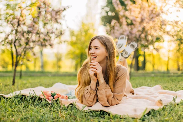 美丽的年轻女子 戴着草帽 戴着一束粉红色的郁金香 坐在开着花的樱花花园里的草坪上 春天开着花的树 — 图库照片