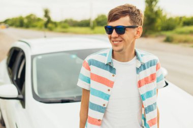 Güneş gözlüklü, yaz elbiseli, arabasının yanında duran, yaz günbatımının tadını çıkaran genç adam, seyahat konsepti.