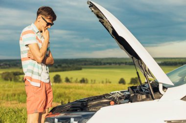 Aracın arızası. Yoğunlaşmış genç bir adam bir arabanın kaputuna tutunup güneşli bir günde dışarıda dikilirken içeriye bakıyor.
