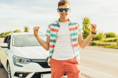 Güneş gözlüklü, arabasının yanında duran mutlu genç adam, güneşli bir yaz gününde, kaslarını açıp, bir güç ve başarı hareketi gösteriyor.