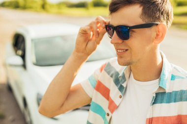 Güneş gözlüklü mutlu genç adam, arabasının yanında duruyor, güneşli bir yaz gününde yolda, yaz tatili konsepti.