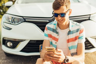 Güneş gözlüklü mutlu genç adam, güneşli bir yaz gününde arabasının yanında yolda otururken cep telefonu kullanıyor.