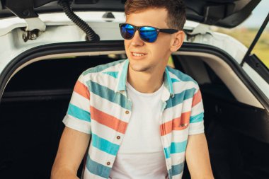 Güneş gözlüklü genç adam, bir arabanın bagajında oturuyor, yolda, gezintiye hayran, yaz tatili, turizm, seyahat