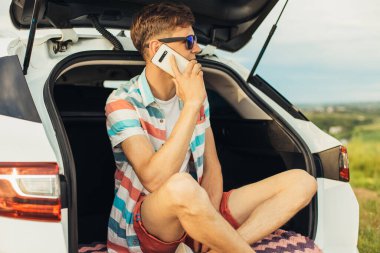 Güneş gözlüklü genç adam bir arabanın bagajında oturuyor ve cep telefonuyla konuşuyor, yaz tatili, turizm, seyahat...