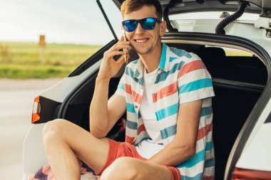 Güneş gözlüklü genç adam bir arabanın bagajında oturuyor ve cep telefonuyla konuşuyor, yaz tatili, turizm, seyahat...