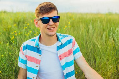 Kırsalda, yeşil tarlada ya da çayırda, tişört giymiş, gülümseyen genç bir adam.