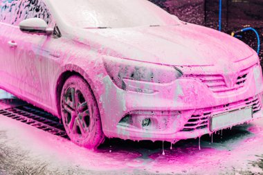 Araba yıkama konsepti, pembe köpükle kaplı beyaz spor araba ön görünümü, profesyonel temizlik ve araba yıkama.