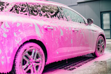 Araba yıkama konsepti, pembe köpükle kaplı beyaz spor araba ön görünümü, profesyonel temizlik ve araba yıkama.