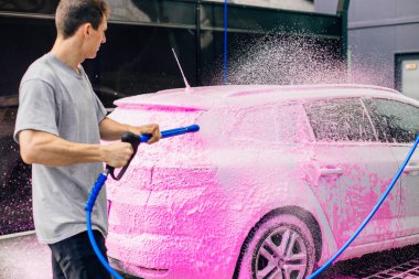 Araba yıkama konsepti, pembe köpükle kaplı beyaz spor araba ön görüş, profesyonel temizlik ve araba yıkama, yüksek basınçlı suyla araba yıkayan adam.