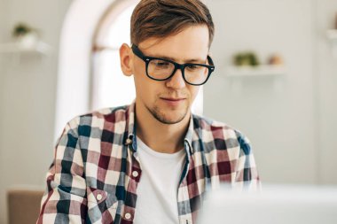 Genç, gülümseyen bir iş adamı, dizüstü bilgisayar kullanıyor, internette çalışıyor, bir kafede modern bir dizüstü bilgisayarla, kitaplarla dolu bir duvarın arkasında oturuyor.