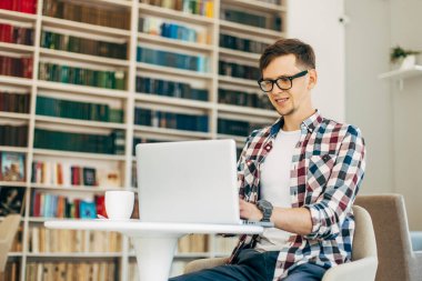 Gözlük takan ve gülümseyen mutlu genç adam, dizüstü bilgisayarıyla çalışıyor, kütüphanede kitaplarla, masada otururken bir fincan kahveyle.
