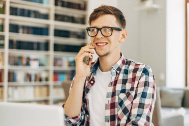 Gülümseyen başarılı erkek öğrenci, gözlüklü, kitaplarla dolu bir kütüphanede cep telefonuyla konuşan.