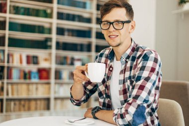 Gözlüklü ve ekose gömlekli genç bir öğrenci kahve içiyor ve üniversite kütüphanesinde arka planda kitaplarla oturuyor..