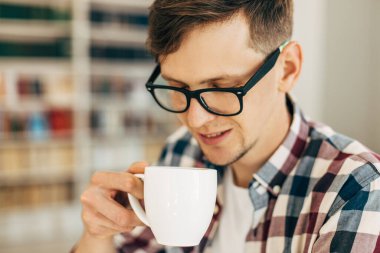 Gözlüklü ve ekose gömlekli genç bir öğrenci kahve içiyor ve üniversite kütüphanesinde arka planda kitaplarla oturuyor..