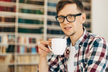 Gözlüklü ve ekose gömlekli genç bir öğrenci kahve içiyor ve üniversite kütüphanesinde arka planda kitaplarla oturuyor..