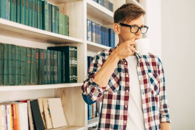 Gözlüklü ve ekose gömlekli genç bir öğrenci kahve içiyor ve üniversite kütüphanesinde arka planda kitaplarla oturuyor..
