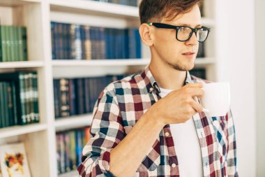 Gözlüklü ve ekose gömlekli genç bir öğrenci kahve içiyor ve üniversite kütüphanesinde arka planda kitaplarla oturuyor..