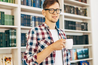 Gözlüklü ve ekose gömlekli genç bir öğrenci kahve içiyor ve üniversite kütüphanesinde arka planda kitaplarla oturuyor..