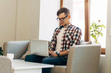 Bilgisayarda çalışan genç bir adam, dizüstü bilgisayar kullanan, evden online çalışma yapan, internette dizüstü bilgisayarda yazan, ekrana bakan, uzaktan kumandayla eğlenen, masada oturan,