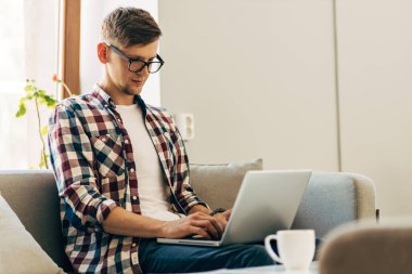 Gözlüklü, dizüstü bilgisayar kullanan, klavyeye metin yazan, e-posta ya da mesaj yazan, sohbet eden, alışveriş yapan, bilgisayarında çalışan başarılı bir serbest çalışan, evdeki koltukta oturan genç adam.