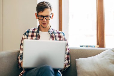 Bilgisayarda çalışan genç bir adam, dizüstü bilgisayar kullanan, evden online çalışma yapan, internette dizüstü bilgisayarda yazan, ekrana bakan, uzaktan kumandayla eğlenen, masada oturan,
