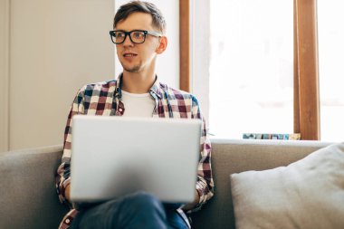 Bilgisayarda çalışan genç bir adam, dizüstü bilgisayar kullanan, evden online çalışma yapan, internette dizüstü bilgisayarda yazan, ekrana bakan, uzaktan kumandayla eğlenen, masada oturan,