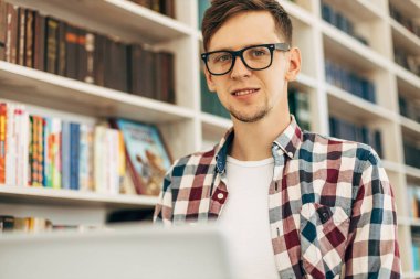 Genç gülümseyen öğrenci sınava hazırlanırken ders kitabı hakkında notlar alıyor. Ödev yapan adam çalışma masasında modern dizüstü bilgisayarla oturuyor.