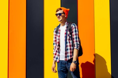 Kırmızı şapkalı ve güneş gözlüklü yakışıklı bir hippinin şehir portresi, şık bir ekoseli gömlek, izole edilmiş renkli bir arka plan.