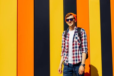 Kırmızı şapkalı ve güneş gözlüklü yakışıklı bir hippinin şehir portresi, şık bir ekoseli gömlek, izole edilmiş renkli bir arka plan.