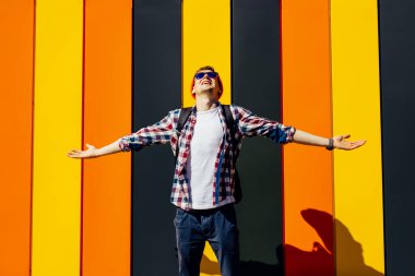 Mutlu özgür genç hippi, gözlüklü ve şapkalı, elleri havada, gökyüzüne bakıyor, şehrin arka planında, özgürlük ve seyahat konseptinde