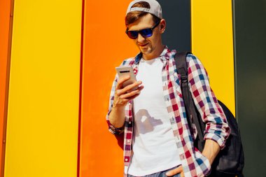 Güneş gözlüklü ve şapkalı genç bir adam şehirde renkli bir duvara yaslanmış bir cep telefonu kullanıyor.