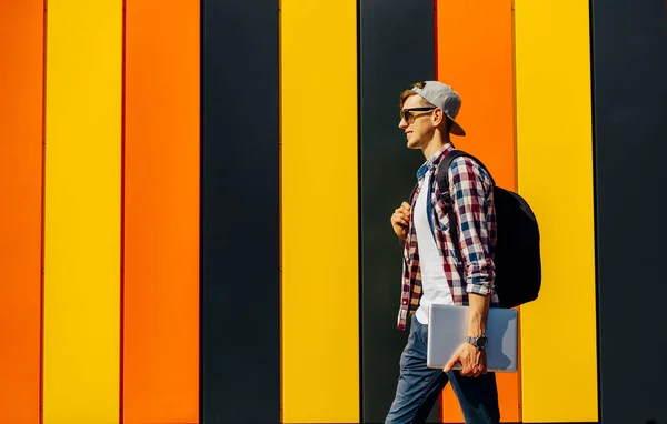 Stylish young man with a briefcase, in sunglasses and a cap, walks along a city street with a laptop, against the background of a colorful wall, the concept of success, business