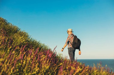 Genç adam, gömlek ve gözlüklü, bir sırt çantasıyla yürüyüşe çıkıyor, gülümseyen adam yokuş yukarı çıkıyor, macera dolu bir hayat sürüyor, yaz tatili vahşi doğada tek başına.