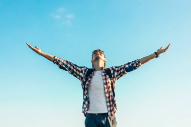 Sırt çantalı genç adam dağın tepesinde duruyor, mavi gökyüzünün arka planında, vadinin manzarasının tadını çıkarıyor.