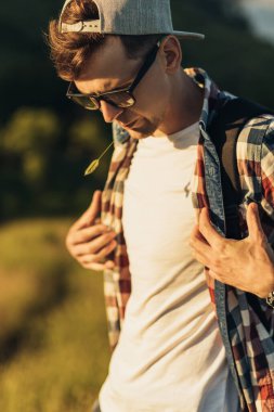 Genç ve yakışıklı bir adamın portresi, gözlüklü ve şapkalı, gülümseyen genç bir adam, güzel bir ormanın arka planında, sokakta dikiliyor.