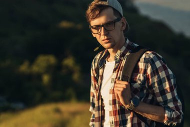 Genç ve yakışıklı bir adamın portresi, gözlüklü ve şapkalı, gülümseyen genç bir adam, güzel bir ormanın arka planında, sokakta dikiliyor.