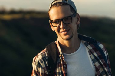 Genç ve yakışıklı bir adamın portresi, gözlüklü ve şapkalı, gülümseyen genç bir adam, güzel bir ormanın arka planında, sokakta dikiliyor.